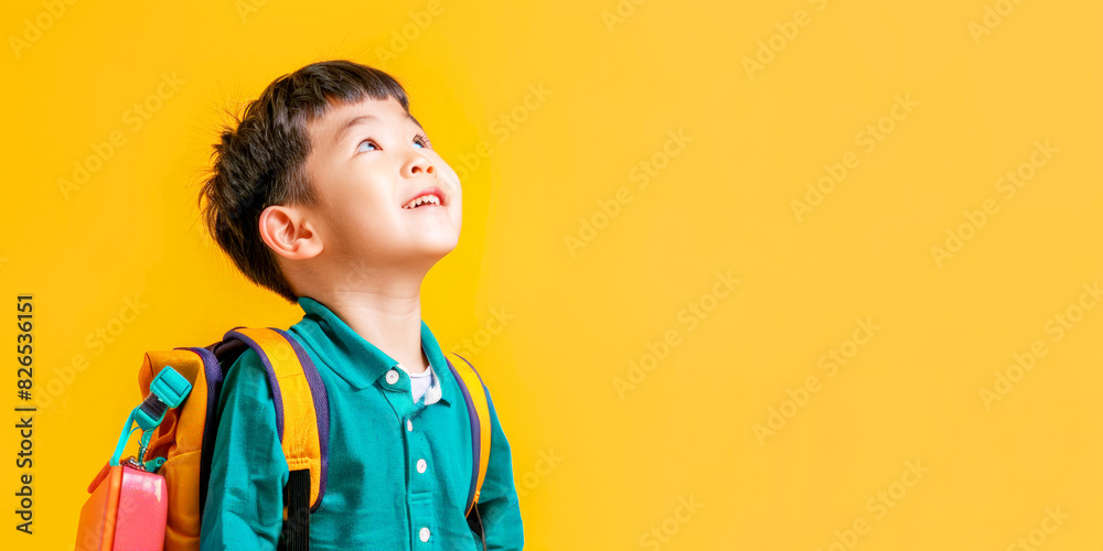 Happy young boy with a colorful backpack looking up, wearing a green shirt, standing against a vibrant yellow background. Happy young boy with a colorful backpack looking up, wearing a green shirt, standing against a vibrant yellow background.