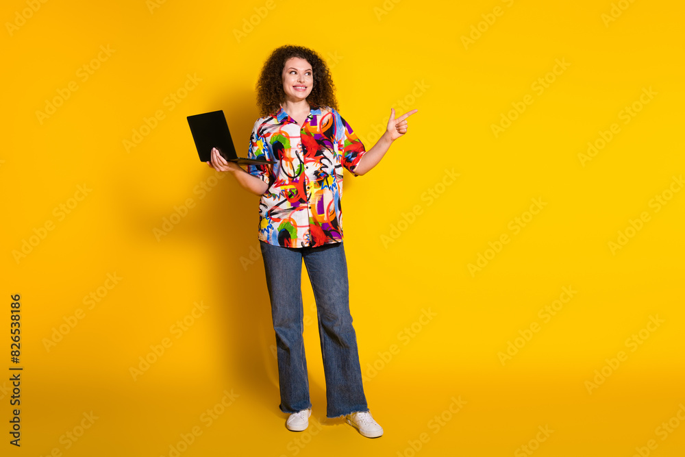 Full body photo of pretty young girl netbook look point empty space wear trendy colorful print outfit isolated on yellow color background Full body photo of pretty young girl netbook look point empty space wear trendy colorful print outfit isolated on yellow color background