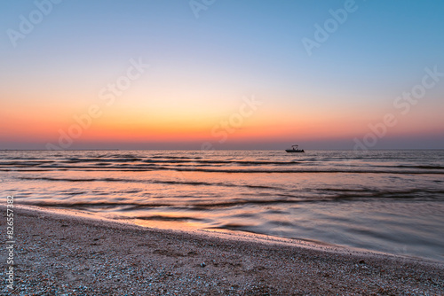 A serene sunset over a calm sea with gentle waves lapping at a pebbled beach. A small boat is silhouetted against the vibrant hues of the sky, creating a tranquil and picturesque scene perfect for con