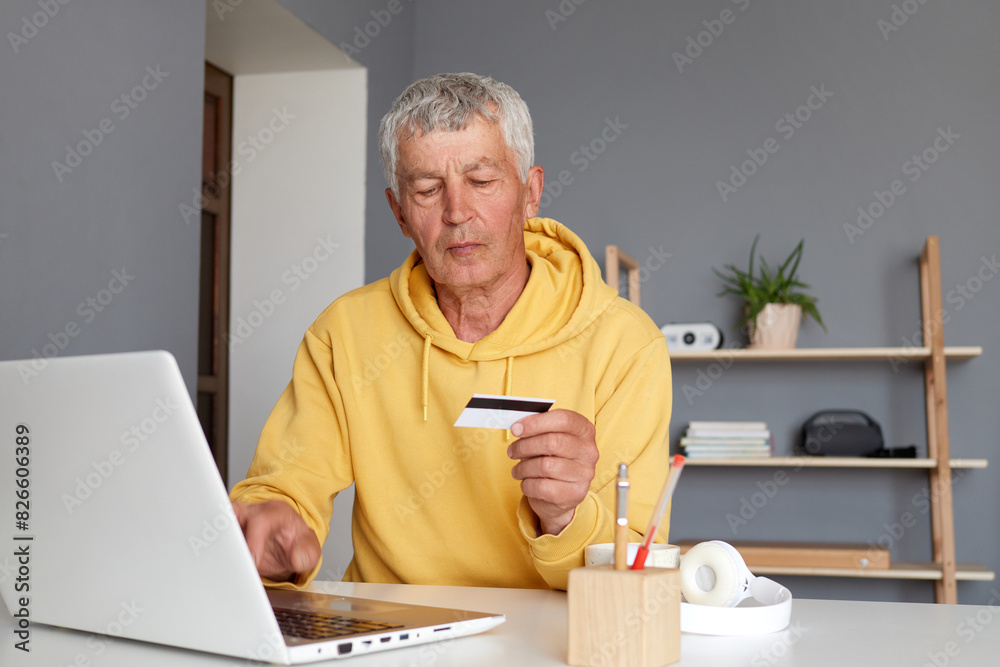 Focused senior man using using credit card to make online payment on laptop holding debit card and using computer for shopping online from home checking credit card Focused senior man using using credit card to make online payment on laptop holding debit card and using computer for shopping online from home checking credit card