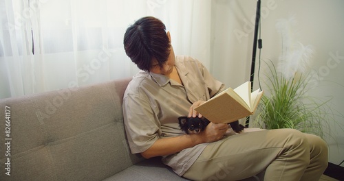 Asian man sitting on a couch stroking lovely fluffy cat with hand on his lap while reading a book in his hand in cozy living room, Relaxed cute animal pet in owner arms, pet lover, leisure hobby