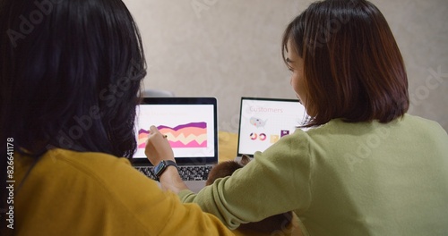 behind view of Two Asian women are working on a presentation together. One of them is pointing at a laptop screen. The laptop screen shows a graph line. The women are discussing the graph