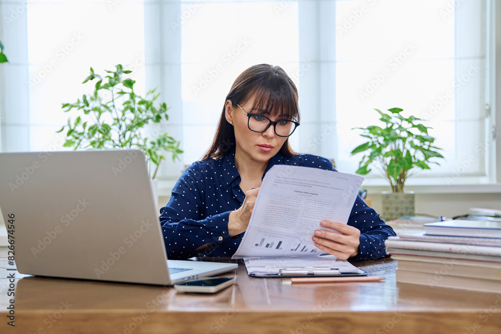 Middle-aged woman working with laptop, business papers, sitting at desk Middle-aged woman working with laptop, business papers, sitting at desk