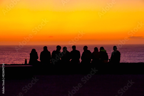 Silhouettes of young people sitting in front of a sea with an orange sunset sky with the sun setting 