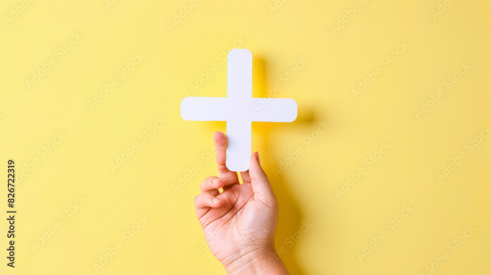 Person's hand holding a cardboard plus sign against a bright yellow background Person's hand holding a cardboard plus sign against a bright yellow background