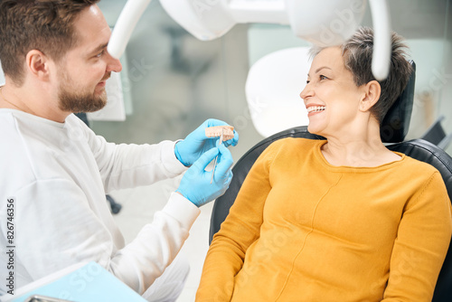 Cheerful aesthetic dentist showing to client how he treat her tooth