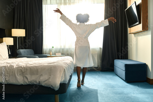 Full length back view of happy young African American woman wearing bathrobe enjoying morning in hotel room and stretching, copy space