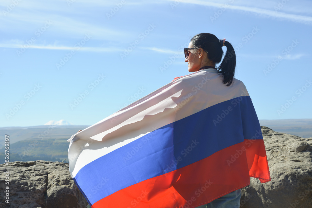 A girl tourist with a Russian flag on her shoulders stands on the top of a mountain and enjoys the beautiful views of the Caucasus Mountains.