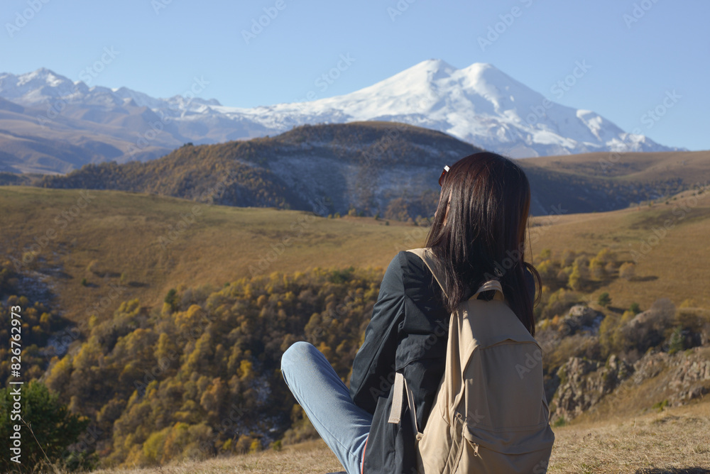 A female traveler with a backpack behind her sits on the grass and enjoys the views of Elbrus. View of Elbrus, North Caucasus, Russia.