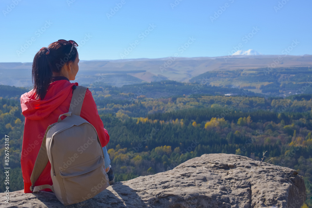A girl tourist with a backpack sits on the top of a mountain and enjoys the beautiful views. A walking woman in an orange jacket rests on a rock, looking at the beautiful sunny landscape of the