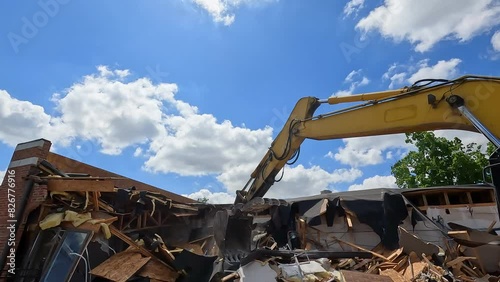 Hydraulic excavator demolishes a building. Industrial equipment with large claw destroys the side of a brick building.  Sunny skies and clouds above the scene. Slow motion views.