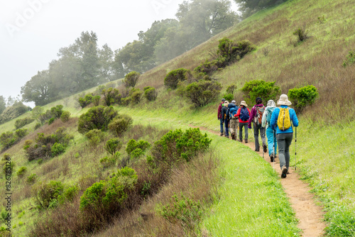 A column of hikers on a trail curving along the side of a slope leading into a fog hidden terrain,  Ipiwa trail, Skyline Ridge OSP, California
