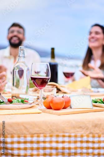 Vertical table foreground. Blurred happy two young adult friends enjoying lunch together outdoors. Cheerful people gathered sitting at table with glass of red wine and food at rooftop summer barbecue 