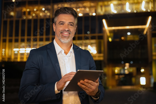 Closeup mature adult professional business man in formal suit working on pc computer. Smiling businessman ceo holding digital tablet using application at office building background in evening outdoors