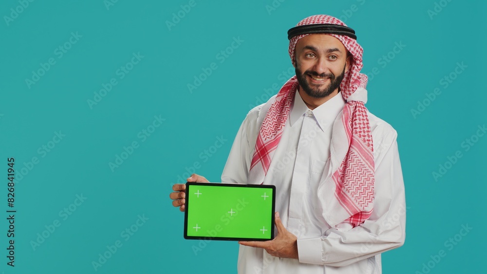 Arab man showing greenscreen on tablet over blue background, presenting isolated copyspace mockup template. Young adult in islamic clothes holding device with blank chromakey display. Arab man showing greenscreen on tablet over blue background, presenting isolated copyspace mockup template. Young adult in islamic clothes holding device with blank chromakey display.