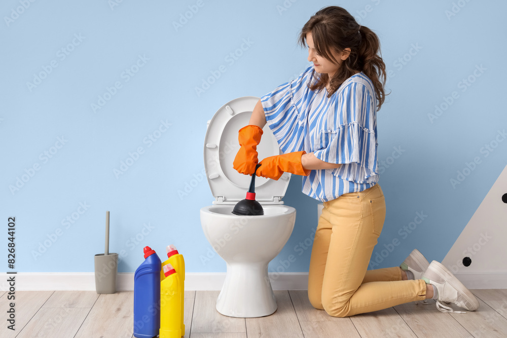 Young woman cleaning toilet bowl with plunger near blue wall Young woman cleaning toilet bowl with plunger near blue wall