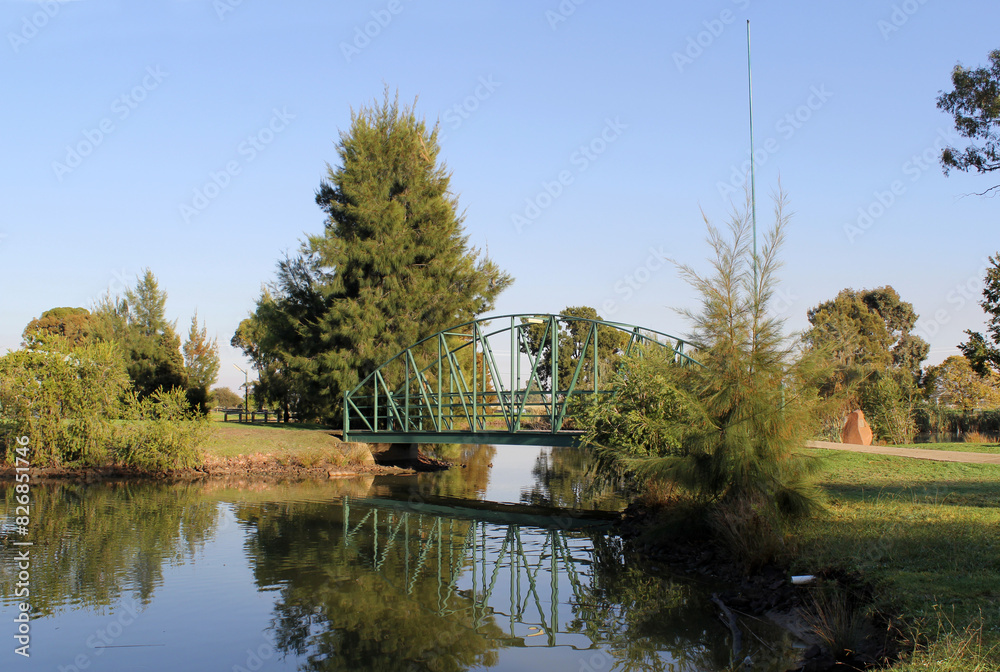 Green metal bridge over a lake of water surrounded by trees and plants at McCaughey Bicentennial Park in Yanco, New South Wales, Australia