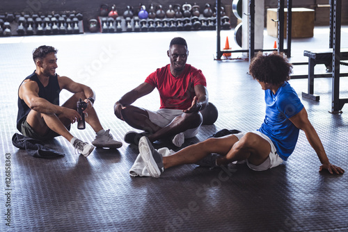 At gym, three young, fit, diverse men sitting on gym floor, chatting and relaxing photo