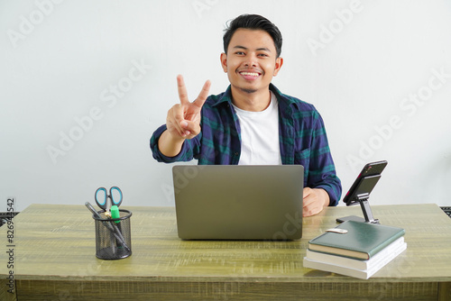 asian man sitting in workplace gesturing peace sign with fingers. Happy guy enjoying remote work at home. Concept of freelance and technology.