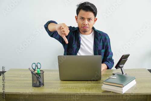 Displeased young asian businessman sitting office workplace showing thumbs down gesture