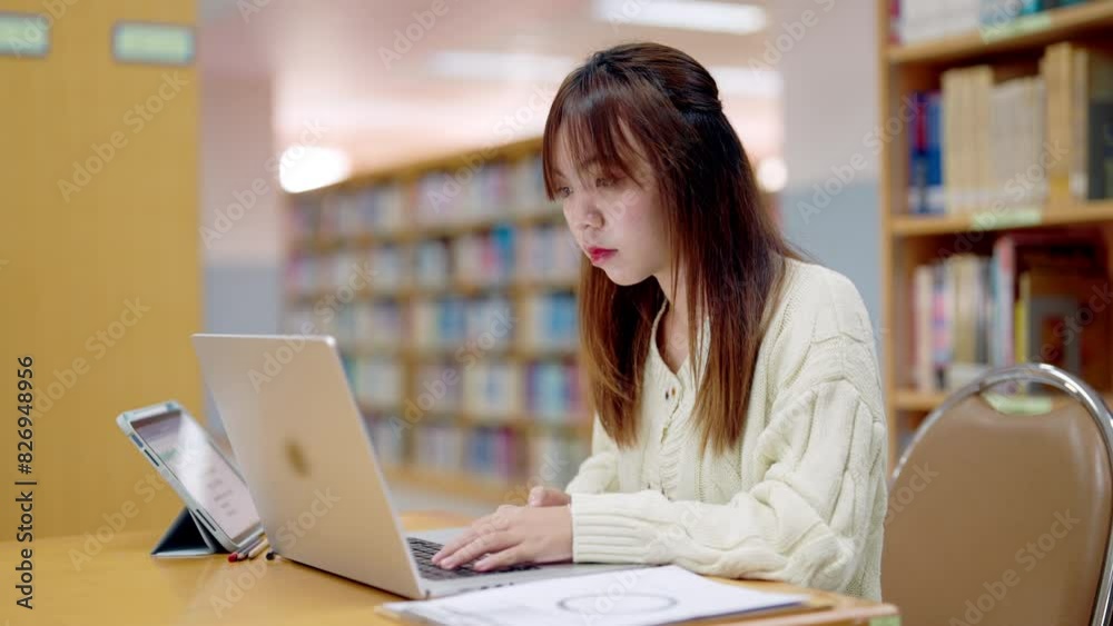 A woman is sitting at a table in a library, typing on a laptop. She is focused on her work, and the scene suggests a quiet and studious atmosphere A woman is sitting at a table in a library, typing on a laptop. She is focused on her work, and the scene suggests a quiet and studious atmosphere