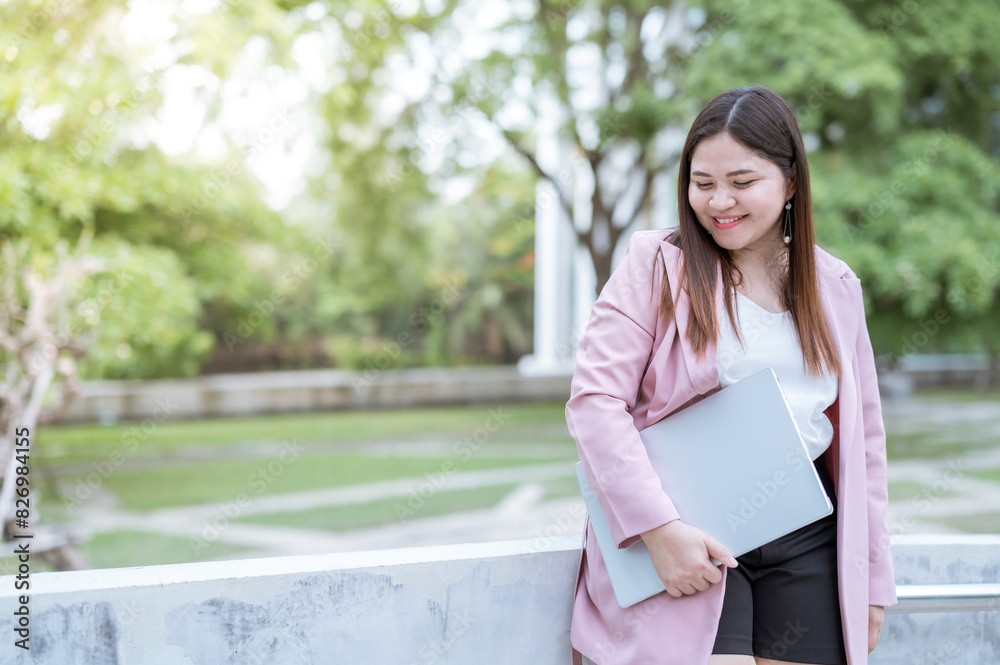 a woman standing outdoors, holding a laptop. She is smiling and looking down. The setting appears to be a park or a campus, The woman is dressed in a casual yet professional outfit, a woman standing outdoors, holding a laptop. She is smiling and looking down. The setting appears to be a park or a campus, The woman is dressed in a casual yet professional outfit,