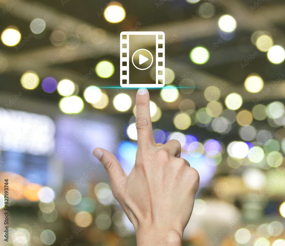 Hand pressing play button with movie flat icon over blur light and shadow of shopping mall, Business cinema online concept Hand pressing play button with movie flat icon over blur light and shadow of shopping mall, Business cinema online concept
