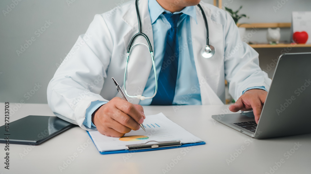 Male doctor sitting at work desk checking work plans and writing document reports in hospital office, healthcare worker and doctor service, digital laptop in modern office, medical technology concept. Male doctor sitting at work desk checking work plans and writing document reports in hospital office, healthcare worker and doctor service, digital laptop in modern office, medical technology concept.