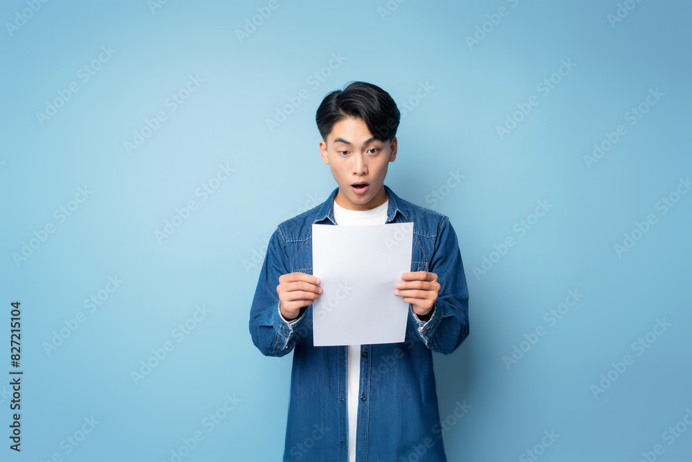Young Asian man has a shocked or surprised expression after reading a piece of paper he's holding in his hands. Isolated on blue background. Young Asian man has a shocked or surprised expression after reading a piece of paper he's holding in his hands. Isolated on blue background.