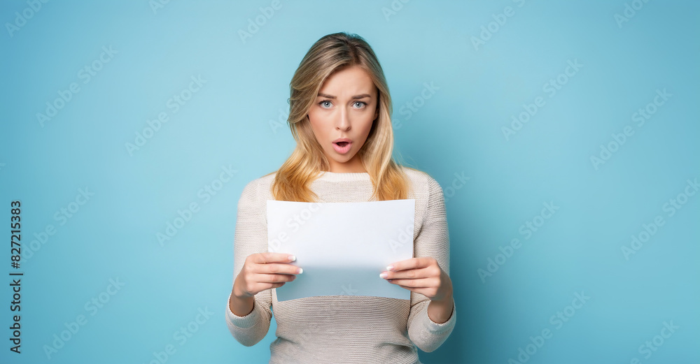 Young blonde woman, holding a piece of paper in her hands, with shocked or surprised expression. Received bad news or n expensive bill. Isolated on blue background. Young blonde woman, holding a piece of paper in her hands, with shocked or surprised expression. Received bad news or n expensive bill. Isolated on blue background.