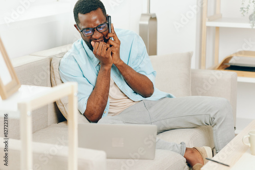 Smiling African American Man using Laptop and Phone on Sofa in Home Office for Online Shopping and Communication