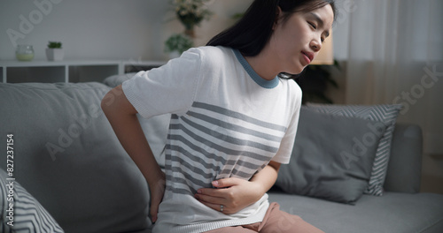 Portrait of Young asian woman has waist pain or lumbago and massaging her loin while sitting on sofa in the living room, healthcare woes photo