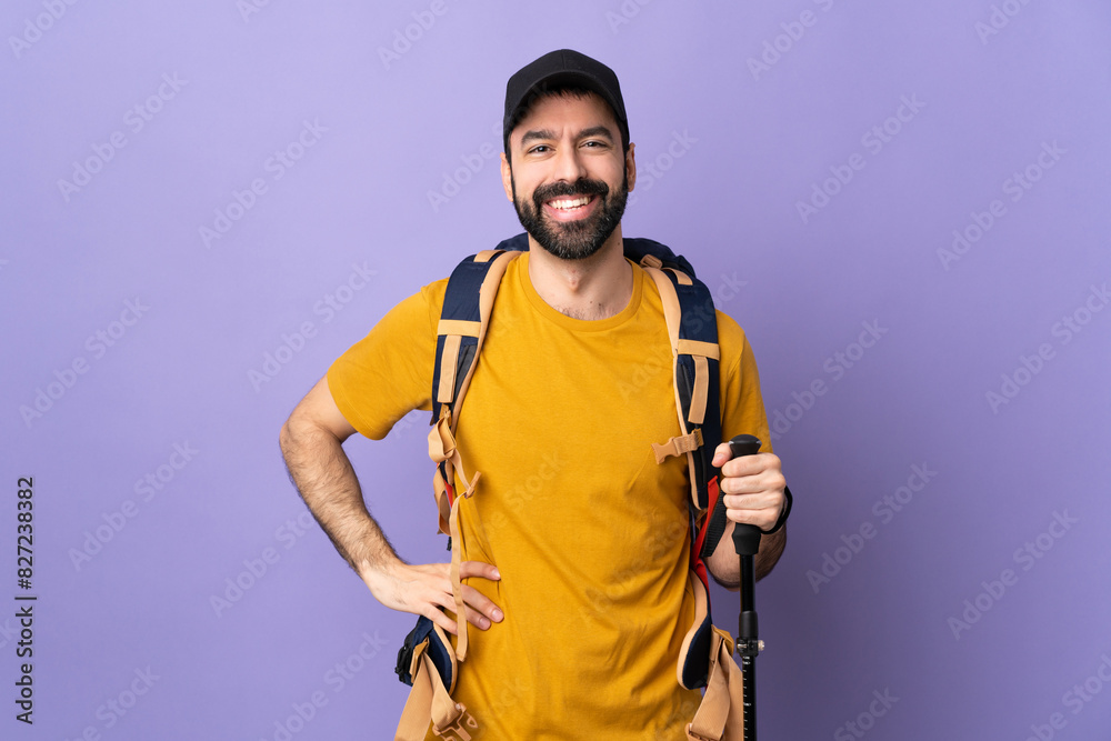 Caucasian handsome man with backpack and trekking poles over isolated background posing with arms at hip and smiling Caucasian handsome man with backpack and trekking poles over isolated background posing with arms at hip and smiling