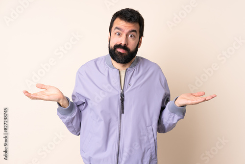 Caucasian man with beard wearing a jacket over isolated background having doubts while raising hands photo