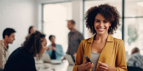 Professional black woman using a smart phone in an office. Happy business woman smiling at the camera in a boardroom. Colleagues having a meeting in the background