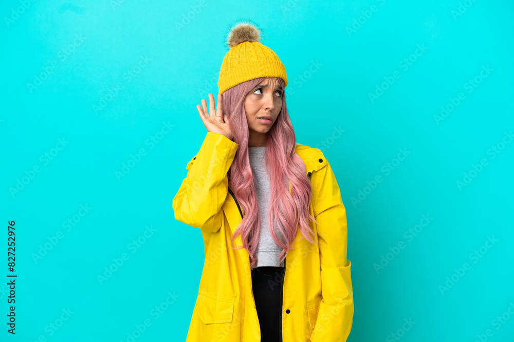 Young woman with pink hair wearing a rainproof coat isolated on blue background listening to something by putting hand on the ear Young woman with pink hair wearing a rainproof coat isolated on blue background listening to something by putting hand on the ear