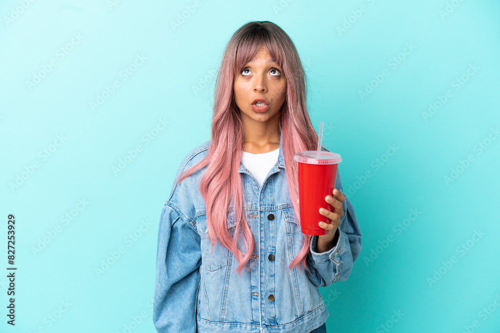 Young mixed race woman drinking a fresh drink isolated on blue background looking up and with surprised expression Young mixed race woman drinking a fresh drink isolated on blue background looking up and with surprised expression