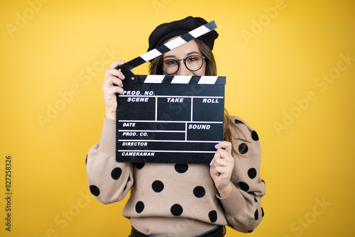 Young beautiful brunette woman wearing french beret and glasses over yellow background holding clapperboard very happy having fun