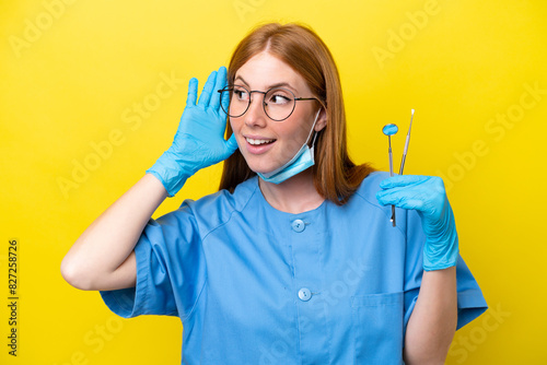 Young redhead Dentist woman isolated on yellow background listening to something by putting hand on the ear photo