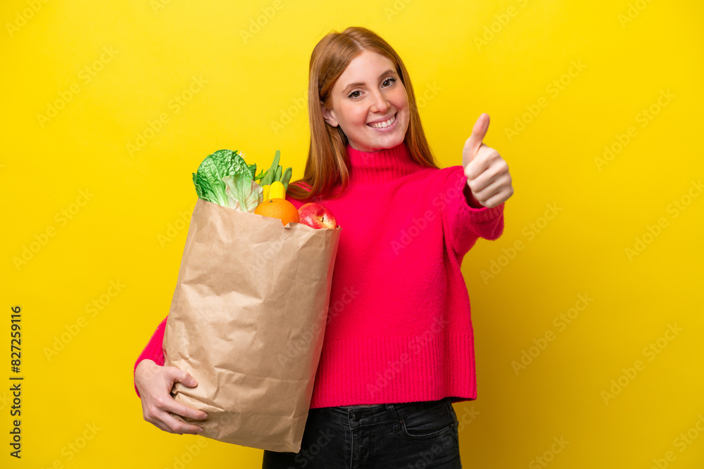 Young redhead woman holding a grocery shopping bag isolated on yellow background with thumbs up because something good has happened Young redhead woman holding a grocery shopping bag isolated on yellow background with thumbs up because something good has happened