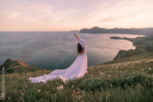 A woman in a white dress stands on a grassy hill overlooking the ocean