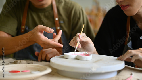 A close-up of Two students painting ceramic heart-shaped pieces with a brush together in an art studio. They work side by side, sharing tools and techniques, fostering creativity and collaboration