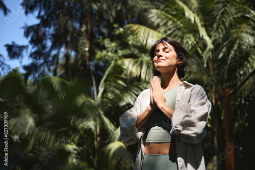 Happy calm young Hispanic woman holding hands in namaste meditating doing yoga breathing exercises with eyes closed feeling peace of mind, mental balance standing in green nature tropical park.