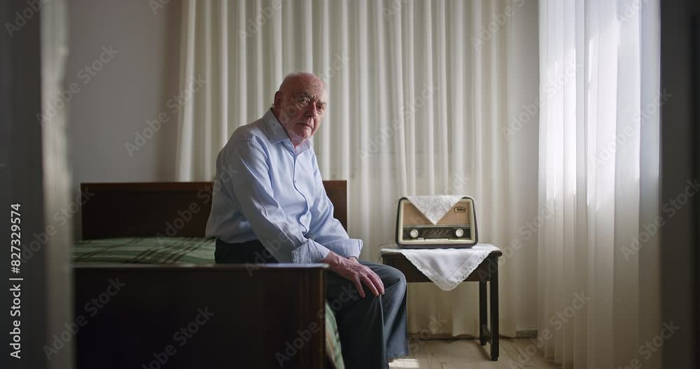An elderly man sits alone on his bed, thoughtfully looking down, in a serene room with a vintage radio on a side table. An elderly man sits alone on his bed, thoughtfully looking down, in a serene room with a vintage radio on a side table.