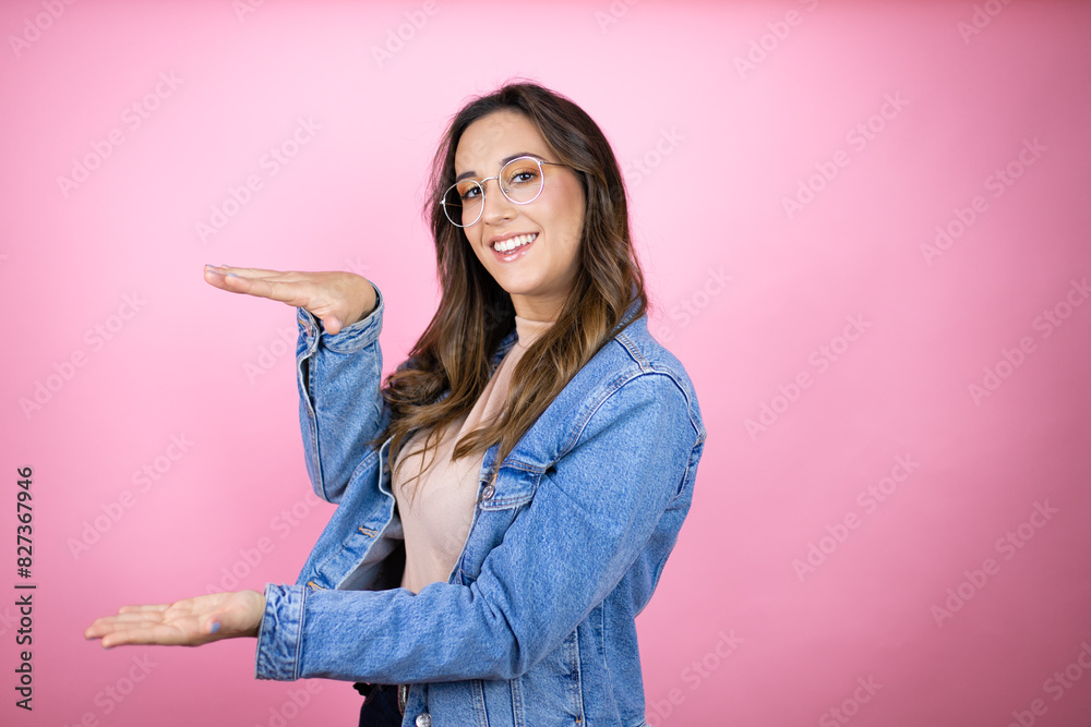 Young beautiful woman wearing denim jacket over isolated pink background gesturing with hands showing big and large size sign, measure symbol. Smiling . Measuring concept. Young beautiful woman wearing denim jacket over isolated pink background gesturing with hands showing big and large size sign, measure symbol. Smiling . Measuring concept.