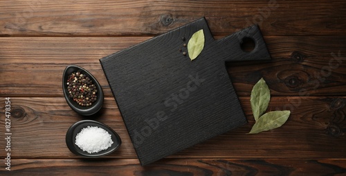 Black cutting board, salt, pepper and bay leaves on wooden table, flat lay. Space for text photo