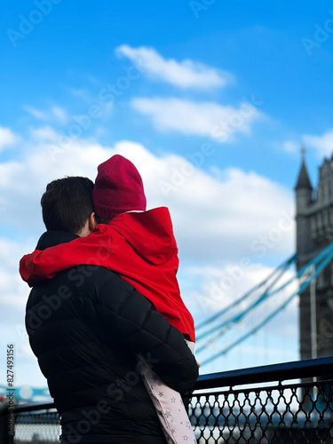 father holds his child, both in warm clothes and look at Tower Bridge in London under blue sky. family travel concept