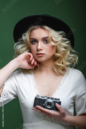 Pretty blonde girl with modern makeup and stylish hairstyle in blackt fedora hat posing with a camera in her hand on a green background. Fashion and beaty. Curly beautiful hair photo