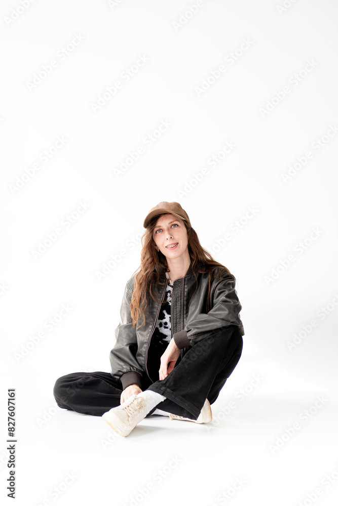 Young woman in a bomber jacket, jeans and a cap with braces poses while sitting in the studio, on a white background Young woman in a bomber jacket, jeans and a cap with braces poses while sitting in the studio, on a white background