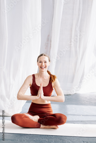A slender woman with braces in a brown sports uniform sits in the lotus position against a background of white curtains and meditates. Yoga classes in the loft studio